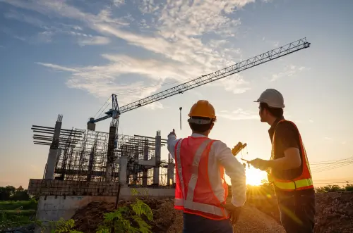 Two men in PPE looking at the bones of a building, sunet in the background